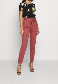 Blouse noire à fleurs avec manches courtes, associée à un pantalon rose taille haute avec ceinture et coupe fuselée. Talons noirs à brides.