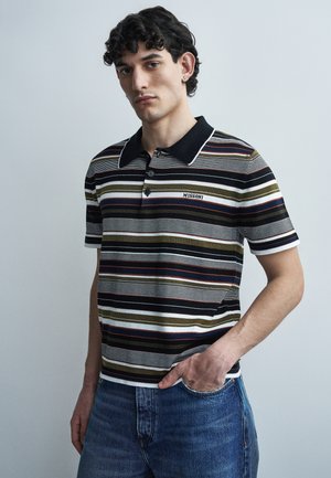 Νέος άνδρας με σγουρά μαλλιά που φοράει ριγέ πόλο μπλούζα με το λογότυπο "MISSONI" και μπλε τζιν, ποζάρει με το ένα χέρι στην τσέπη μπροστά σε φωτεινό φόντο.