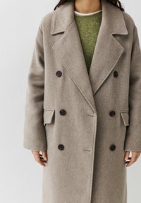 Persoon draagt een taupekleurige wollen mantel met dubbele rij knopen over een groene trui en een wit ondershirt, staand tegen een effen achtergrond.