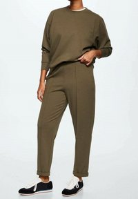 Sweat-shirt vert olive et pantalon fuselé assorti, tous deux en tissu doux. Le mannequin porte des baskets noires et blanches avec des accents de semelle clairs.