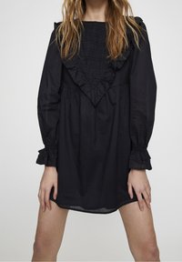 Robe noire en tissu léger, présentant un corsage froncé, un col à volants et des manches longues avec des accents de volants aux poignets.