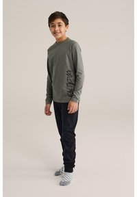 Olívazöld, hosszú ujjú ing sötét felirattal az oldalán, fekete nadrággal és szürke kockás zoknival párosítva, egy fiú által viselve.