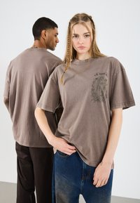 Ruda per didelį T-shirt marškinėlį iš švelnaus medvilnės mišinio, su spausdinta dizaino dekoracija ant priekio. Trumpomis rankovėmis ir laisvu siluetu, derinama su tamsiomis ir mėlynomis džinsomis.