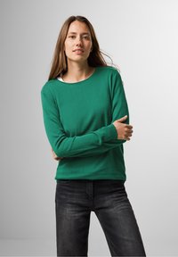 Grüner, gestrickter Pullover mit rundem Halsausschnitt und langen Ärmeln, kombiniert mit dunklen Jeans. Glatte Textur mit enganliegender Silhouette.