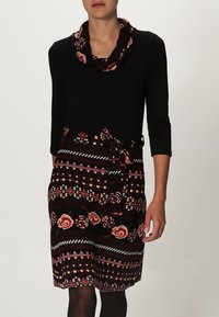 Maglia nera a maniche lunghe con collo a caramella, abbinata a una gonna a fantasia con motivi floreali e geometrici nei toni del bordeaux, crema e verde scuro.