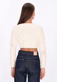 Gekürzter cremefarbener Strickpullover mit bunten Sprenkeln, gerippten Bündchen und detaillierten offenen Strickmustern an den Ärmeln, kombiniert mit dunkelblauen Jeans.