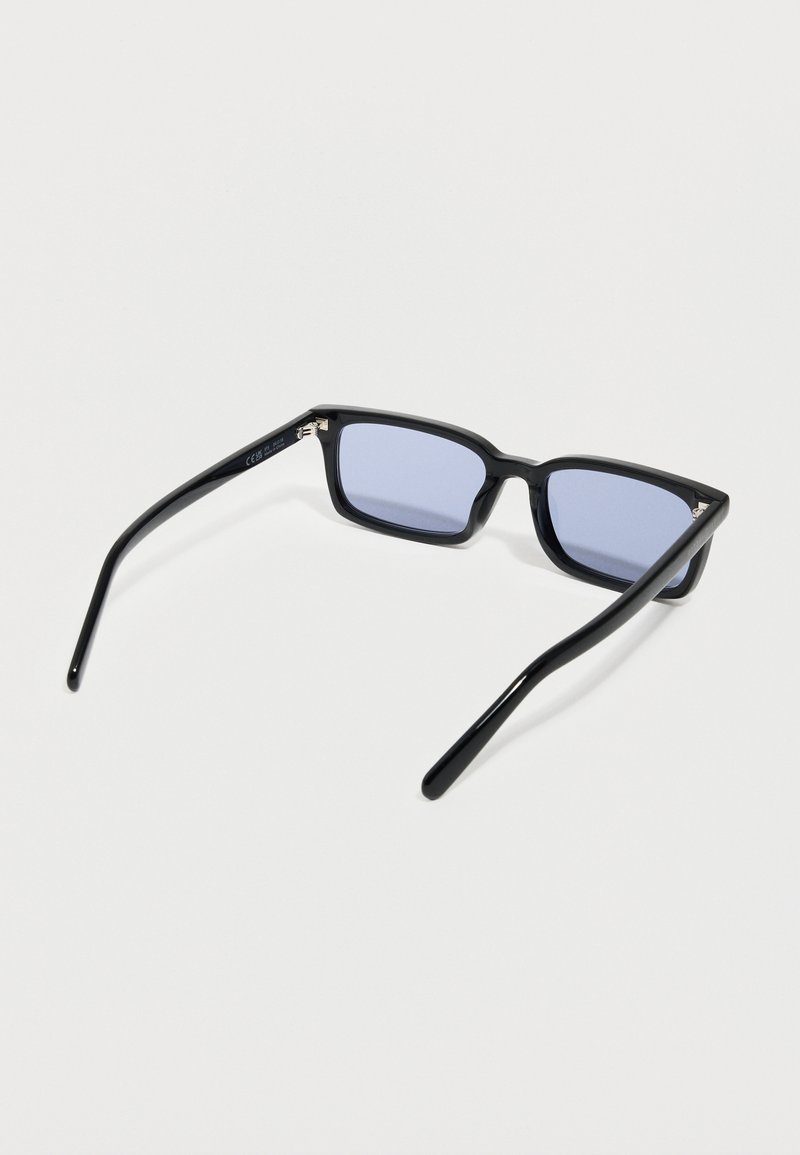 Lunettes de soleil rectangulaires noires avec des branches fines et des verres teintés en bleu clair reposant sur une surface blanche.