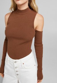 Brauner, gerippter Turtleneck-Bodysuit mit kaltem Schulterdesign und langen Ärmeln, kombiniert mit hoch geschnittenen weißen Jeans mit Knopfdetails.