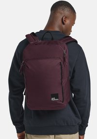 Zaino in tessuto bordeaux con una trama liscia, numerosi scomparti con zip e dettagli neri, caratterizzato da un logo sulla parte anteriore inferiore.