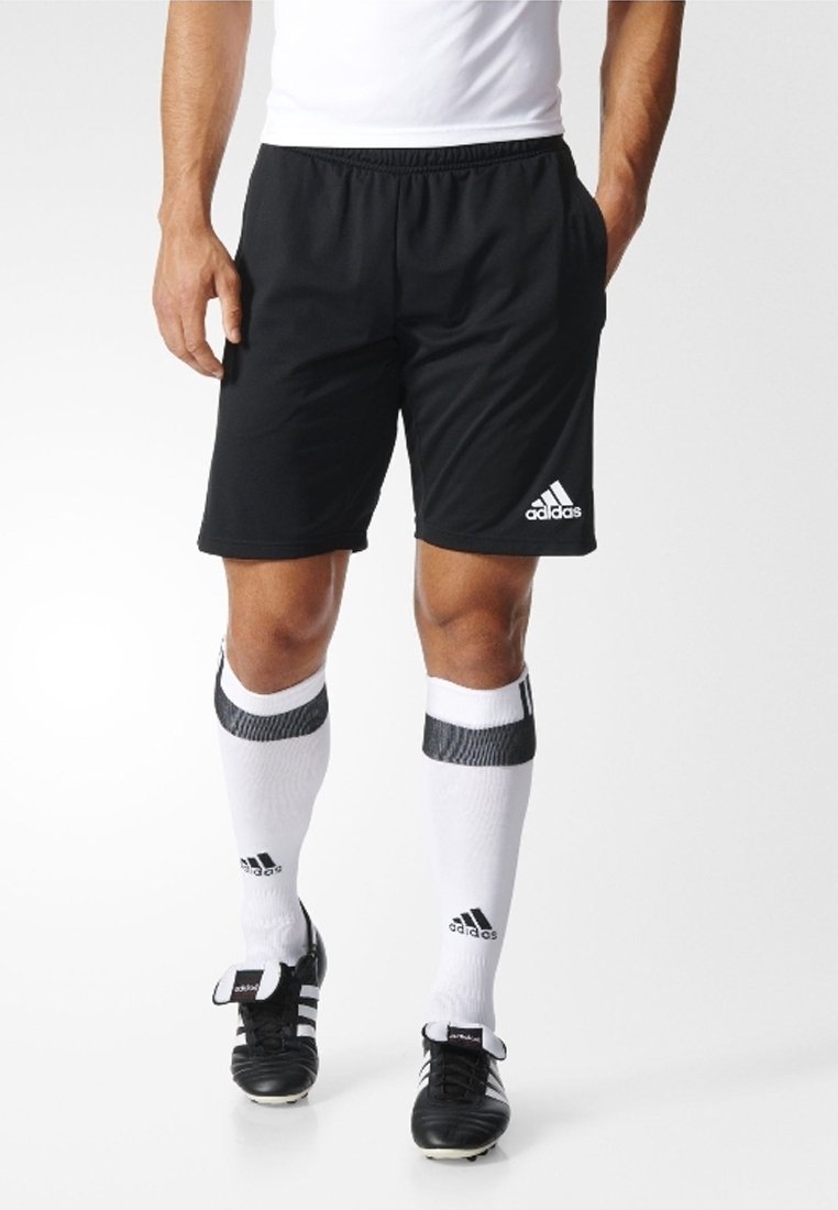 Muž oblečený v černých šortkách Adidas, bílých ponožkách do půli lýtek Adidas s černými pruhy a černých fotbalových kopačkách Adidas, stojící na bílém pozadí.