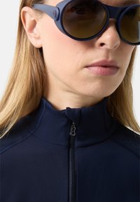 Detailní záběr osoby nosící tmavě modré sluneční brýle a námořnicky modrý zip-up top s prošitými detaily na ramenou a límci.