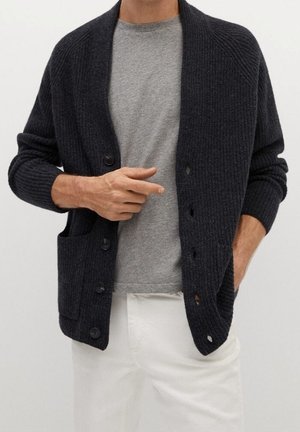 Homme portant un cardigan en maille côtelée gris foncé sur un t-shirt gris clair et un pantalon blanc, une main dans la poche et l'autre ajustant le cardigan.