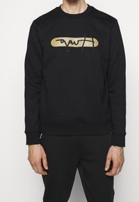 Uomo che indossa una felpa nera con un ovale beige e un design astratto di scritte nere al centro, abbinata a pantaloni neri.