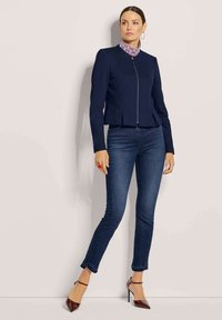 marineblaue, taillierte Jacke mit einem Reißverschluss und einem gemusterten Kragen. Kombiniert mit dunklen Skinny-Jeans und burgunderfarbenen spitzen Pumps.