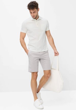 Mann in weißem Poloshirt und hellgrauen Shorts hält eine große weiße Tragetasche, steht mit einer Hand in der Tasche und schaut nach unten.