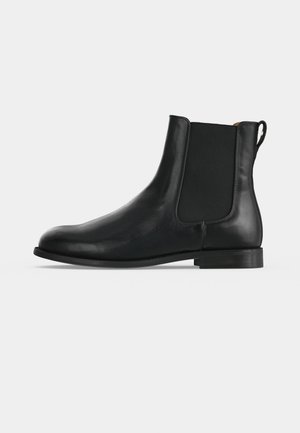Bottine Chelsea en cuir noir avec panneaux latéraux élastiques, petit talon, bout arrondi et languette à l’arrière sur un fond gris uni.
