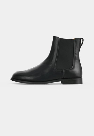 Bottine Chelsea en cuir noir avec panneaux latéraux élastiques, petit talon, bout arrondi et languette à l’arrière sur un fond gris uni.