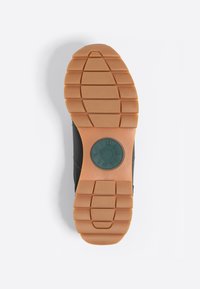 Suela de zapato negra con una suela de goma marrón que presenta un patrón de banda de rodadura horizontal y un acento circular verde con la marca.
