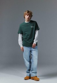 T-shirt en coton vert superposé à un haut blanc à manches longues, associé à un jean bleu clair et des sabots orange, devant un fond neutre.