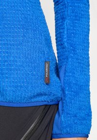 Μπλε φλις ζακέτα με ανάγλυφη επιφάνεια, με λογότυπο Polartec, συνδυασμένη με μαύρο ύφασμα και λεπτομέρεια με φερμουάρ στην τσέπη.