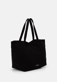 Černá látková taška na rameno s dvojitými uchy a malou obdélníkovou nálepkou blízko spodního okraje na bílém pozadí.