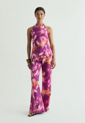 Mujer con top sin mangas de cuello halter y pantalones de pierna ancha con estampado floral en tonos púrpura, rosa y naranja, de pie frente a un fondo claro y liso.