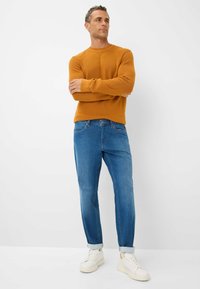 Maglione a maglia senape, jeans in denim blu a gamba affusolata e sneakers bianche. L'outfit presenta linee semplici e una vestibilità rilassata.