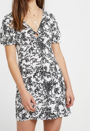 Biała sukienka z krótkim rękawem w czarny liściasty wzór, dekolt w kształcie litery V z ozdobnym pierścieniem z przodu, noszona z warstwowymi złotymi naszyjnikami.