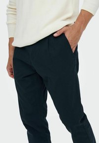 Námořnické kalhoty vyrobené z hladké látky s uvolněným střihem, které mají boční kapsy a jemné přední sklady, kombinované se světle krémovým svetrem.