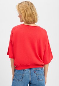 Rode gebreide oversized top met korte mouwen, ronde hals en een zachte textuur, gecombineerd met blauwe denim jeans met achterzakken.