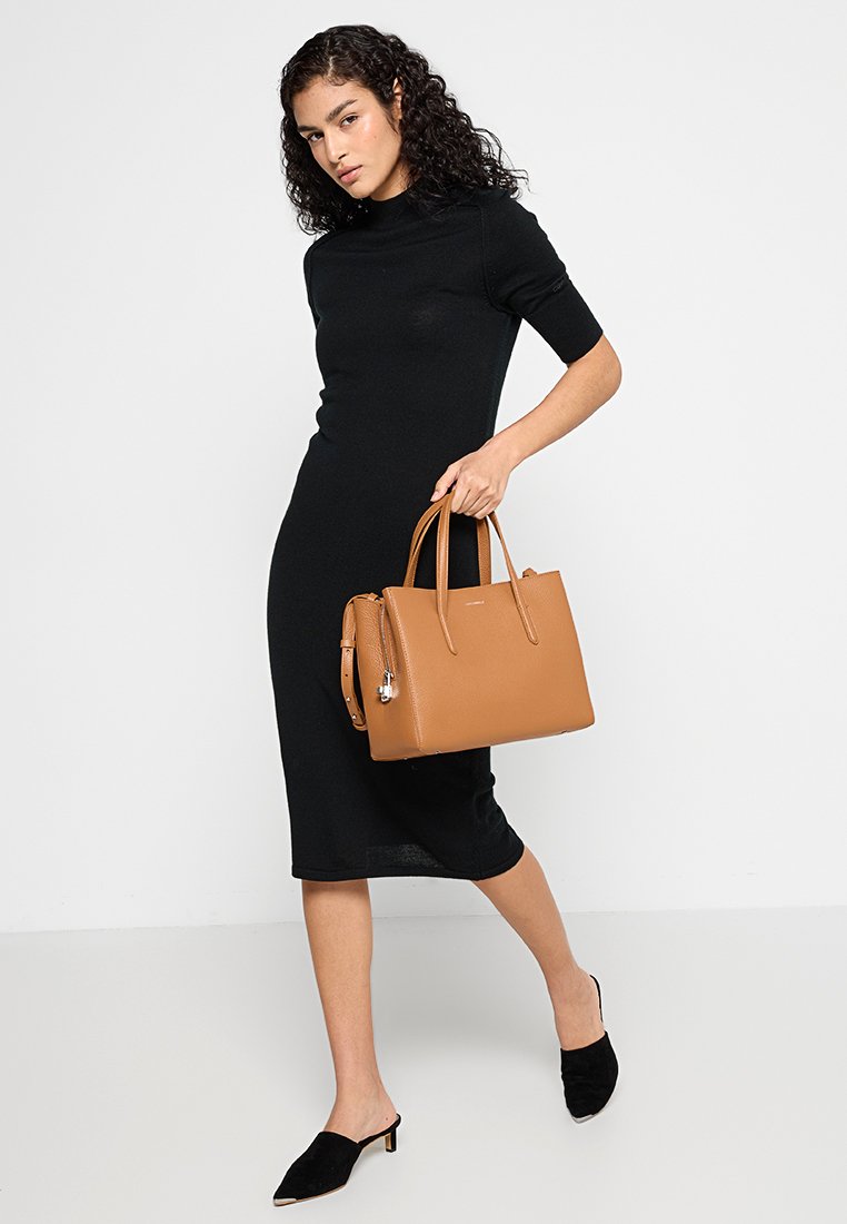 Tanfarbener Lederhandtasche mit doppelten Griffen, die eine schlanke Form und eine strukturierte Oberfläche aufweist. Das Modell trägt ein enges schwarzes Kleid und schwarze Mules.