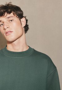 Mørkegrøn crewneck-sweatshirt i tekstureret stof. Har en ribbet krave og detaljerede syninger, båret af en ung mand med krøllet hår.