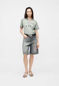 Γυναίκα με ανοιχτό πράσινο t-shirt με την επιγραφή "Property of Diesel", μαύρη ζώνη, γκρι σορτς τζιν Μπέρμουντα και μαύρα σανδάλια με λουράκια, στέκεται μπροστά σε λευκό φόντο.