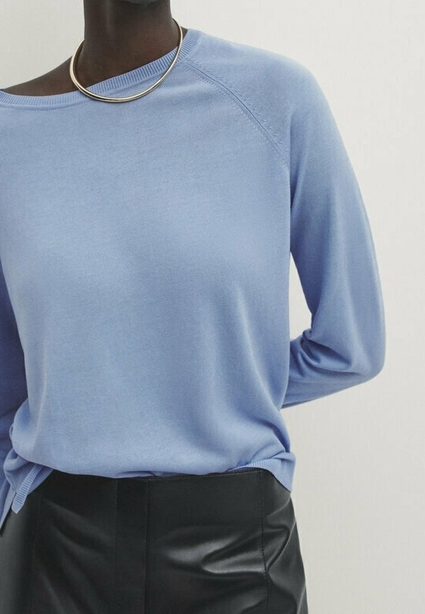 Personne portant un haut à manches longues bleu clair, une jupe noire en cuir et un collier doré, posant devant un fond clair et uni.