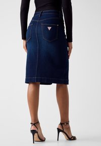 Dunkelblauer Jeansrock mit figurbetonter Silhouette, mittlerer Länge, ausgestattet mit Gesäßtaschen und einem Logoetikett, kombiniert mit schwarzen Riemchenabsätzen.