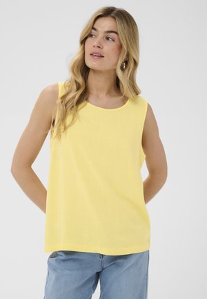 Femeie cu păr lung și blond, purtând un top galben fără mâneci și blugi albastru deschis, uitându-se ușor spre dreapta cu mâinile la spate.