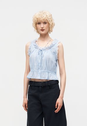 Ξανθό άτομο με κοντά, σγουρά μαλλιά φοράει ανοιχτό μπλε αμάνικο μπλουζάκι με δαντέλα και μαύρο ψηλόμεσο παντελόνι σε λευκό φόντο.