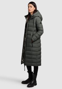 Manteau long, style doudoune, de couleur vert foncé avec une capuche, présentant des sections matelassées horizontales, des poches latérales et une fermeture éclair intégrale. Bottes noires visibles.