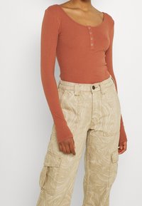 Langärmliger, gerippter Bodysuit in Rostfarbe, mit einem runden Ausschnitt und einer Knopfleiste mit fünf Knöpfen. Kombination mit beigen gemusterten Cargo-Hosen.