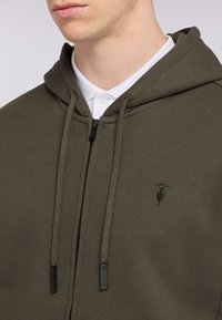 Olivgrön zip-up hoodie med texturerad tyg, huva med dragsnodd och liten broderad logotyp på bröstet. Vit skjorta med krage under.