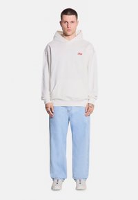 Biała bawełniana bluza z kapturem z kangurzą kieszenią i czerwonym haftowanym logo, w połączeniu z jasnoniebieskimi dżinsami i neutralnymi trampkami.