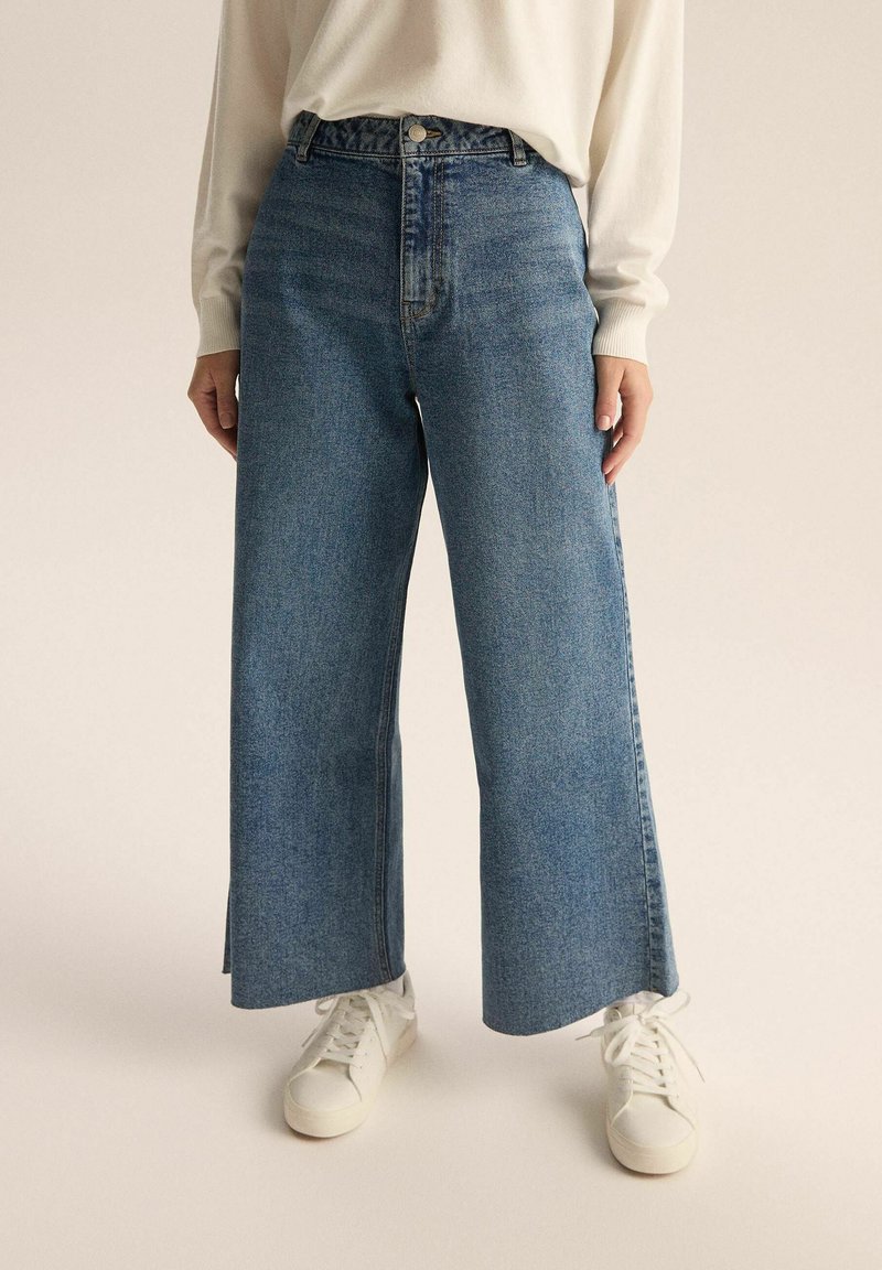 Niebieskie jeansy o szerokich nogawkach i wysokim stanie z detalami w szwie, w połączeniu z białymi sneakersami. Tkanina ma delikatną teksturę.