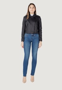 Giacca nera corta con dettagli con zip, abbinata a jeans skinny blu e tacchi beige, su uno sfondo neutro.