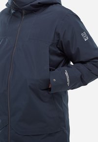 Marineblaue wasserdichte Jacke mit Kapuze, ausgestattet mit Fronttaschen, glatter Textur und Logodetail am Ärmel. Mit Reißverschluss.