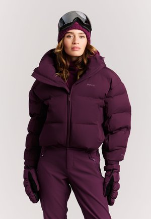 Vrouw draagt paarse skipak, broek, handschoenen en hoed met skibril op haar hoofd tegen een effen achtergrond.