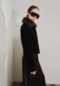 Černá kordurová zkrácená bunda s hnědým kožešinovým límcem, kombinovaná s tmavě hnědou saténovou sukní. Modelka má na sobě velké černé sluneční brýle a náušnice s kamínky.
