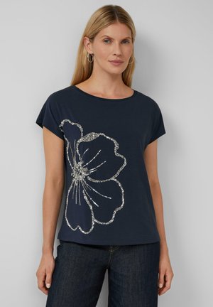 T-shirt bleu marine avec un grand motif floral argenté, manches courtes et col rond, associé à un jean en denim foncé.