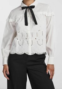 Blusa bianca con colletto a volant e cravatta nera. Presenta un motivo floreale ricamato con ritagli, orlo scallop e maniche lunghe.