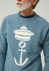 Hombre mayor con barba blanca que lleva un sudadera azul con un gráfico de ancla y boya blanca, acompañado del texto "ALTONADOCK".
