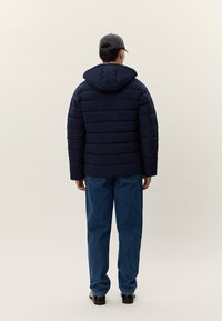 Άνδρας που φοράει μπλε σκούρο μακρύ μπουφάν με κουκούλα, σκούφο, μπλε τζιν και μαύρα παπούτσια, στέκεται μπροστά σε ανοιχτό μπεζ φόντο.